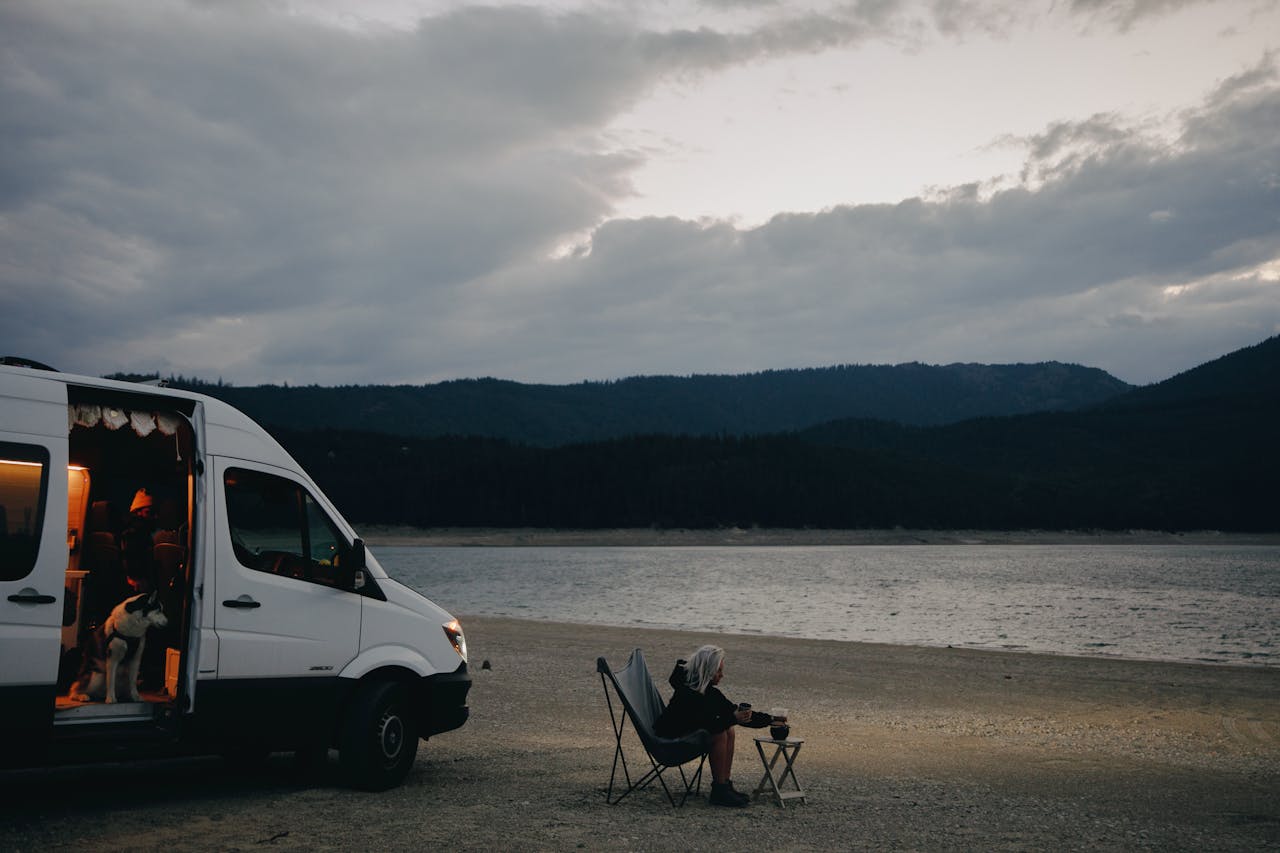 services-02 A woman relaxes by a lakeside, seated next to a campervan with a dog and folding chair under cloudy skies.
